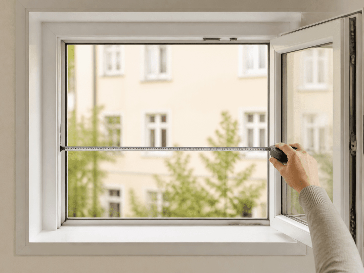 Hand hält ein Maßband quer über die Innenkante eines Fensters